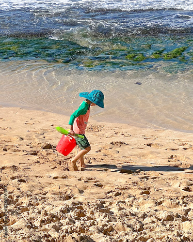 A little girl in colorful clothing,  sporting a turquoise hat, carries a red sand bucket with tools, hunts treasures on beautiful Punaula Beach Ohau. photo