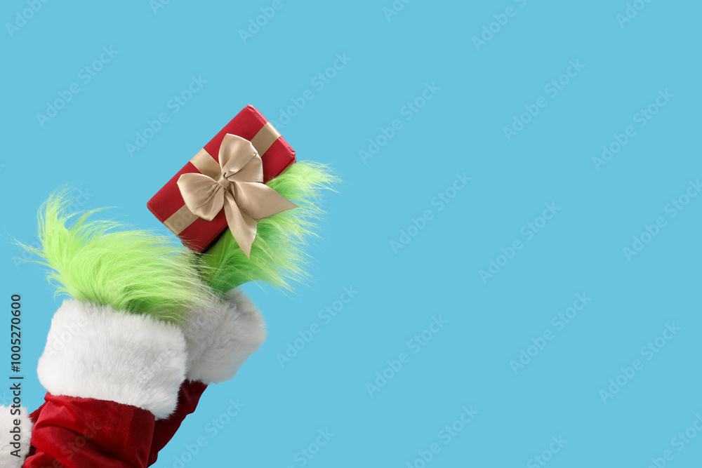 Obraz premium Green hairy hands of creature in Santa costume with Christmas gift box on blue background