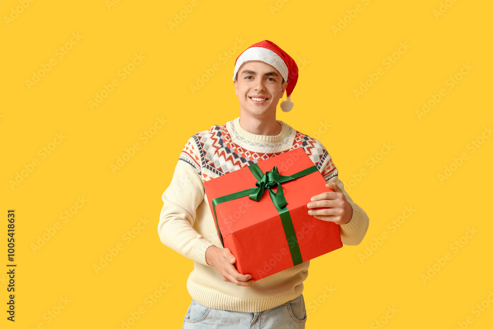 Happy young man in Christmas sweater and Santa hat with gift box on yellow background Happy young man in Christmas sweater and Santa hat with gift box on yellow background