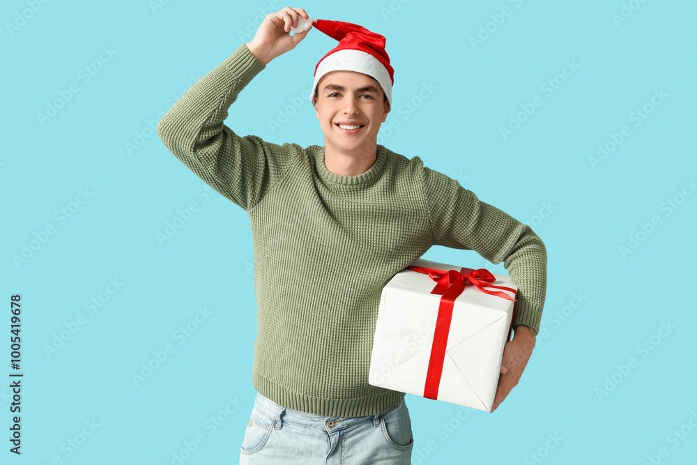 Happy young man in Christmas sweater and Santa hat with gift box on blue background Happy young man in Christmas sweater and Santa hat with gift box on blue background