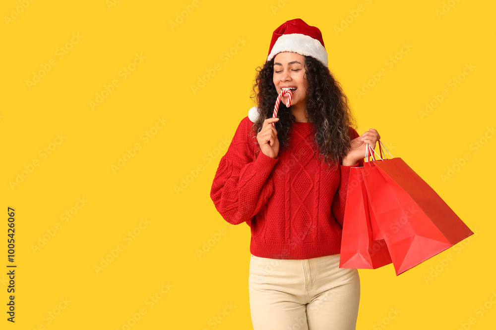 Beautiful young African-American woman in Santa hat with shopping bags and candy cane on yellow background. Christmas celebration Beautiful young African-American woman in Santa hat with shopping bags and candy cane on yellow background. Christmas celebration