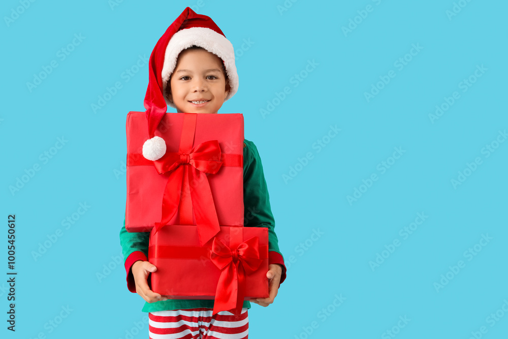 Cute happy little Asian boy in Santa hat dressed as elf with Christmas gift boxes on blue background Cute happy little Asian boy in Santa hat dressed as elf with Christmas gift boxes on blue background