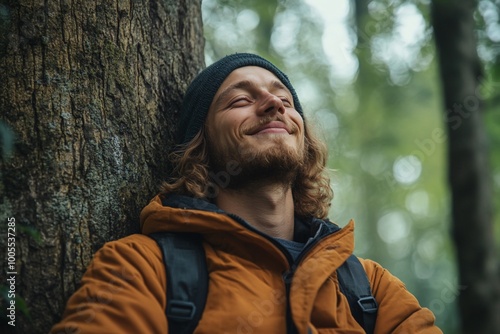 A young man with a beard and beanie smiles while leaning against a tree in the forest.