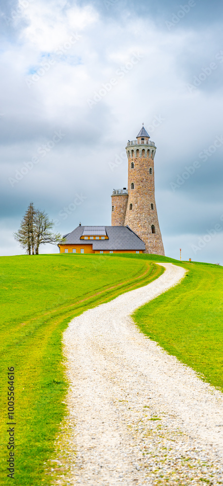 A gravel path leads to the Dalimil Lookout Tower in the Czech Republic. The tower stands tall against a backdrop of lush green grass and a blue sky with fluffy clouds. A gravel path leads to the Dalimil Lookout Tower in the Czech Republic. The tower stands tall against a backdrop of lush green grass and a blue sky with fluffy clouds.