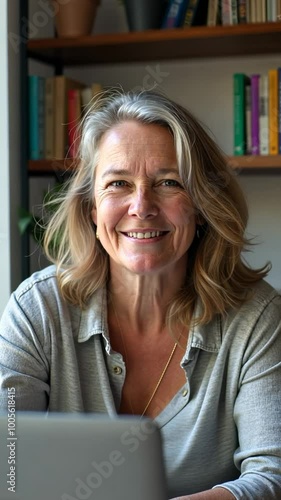 A woman is smiling and sitting in front of a laptop. She is wearing a gray shirt and a necklace