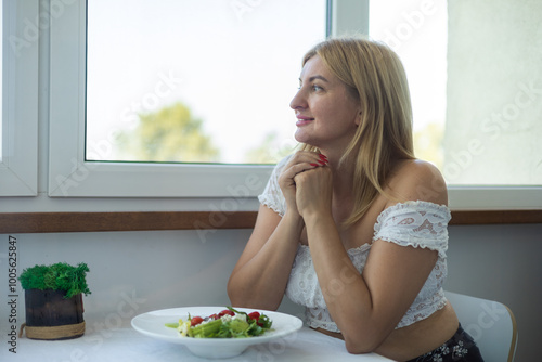 Young happy smiling blonde woman with salad and coffee in kitchen at home