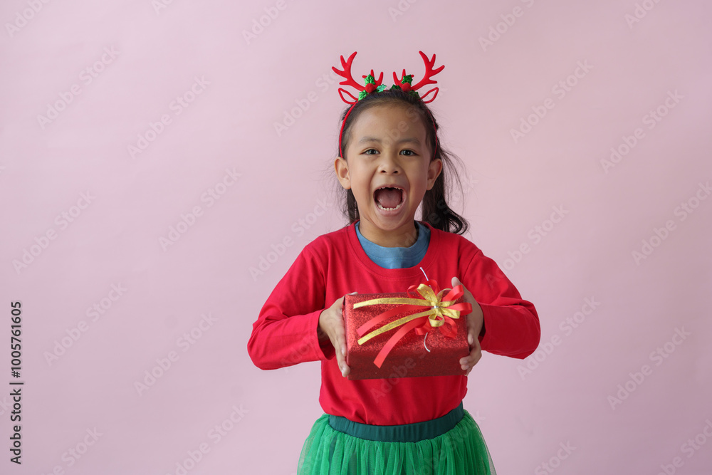 Portrait on a pink background of a cute little girl in a headband holding a happy red gift box with a beautiful green bow in various verses. Christmas Eve New Year and Winter holidays. Portrait on a pink background of a cute little girl in a headband holding a happy red gift box with a beautiful green bow in various verses. Christmas Eve New Year and Winter holidays.