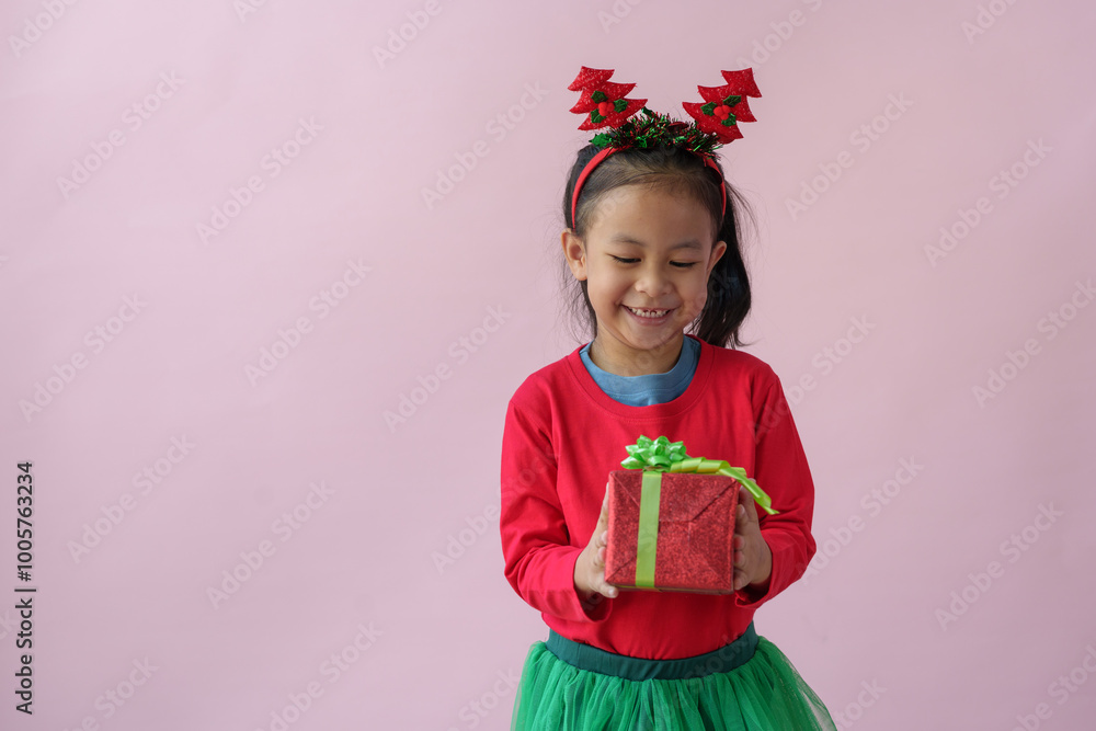 Portrait on a pink background of a cute little girl in a headband holding a happy red gift box with a beautiful green bow in various verses. Christmas Eve New Year and Winter holidays. Portrait on a pink background of a cute little girl in a headband holding a happy red gift box with a beautiful green bow in various verses. Christmas Eve New Year and Winter holidays.