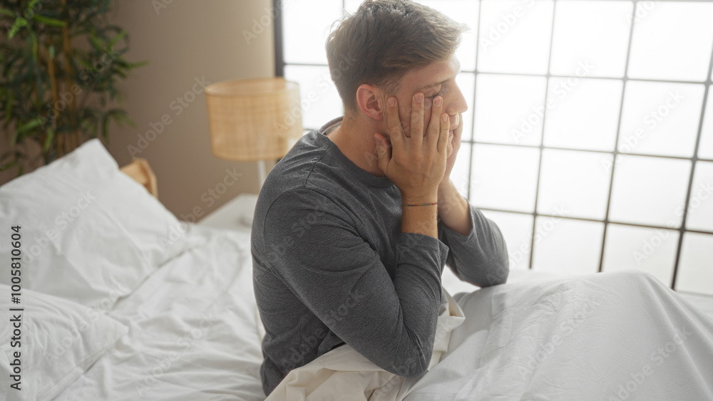 Young man sitting on a bed in a modern bedroom appearing frustrated over a white wall background Young man sitting on a bed in a modern bedroom appearing frustrated over a white wall background
