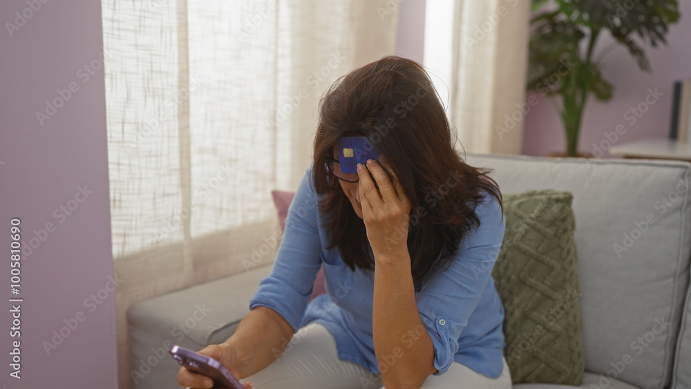 Upset middle-aged woman holding a credit card and smartphone in a living room, suggesting financial distress at home. Upset middle-aged woman holding a credit card and smartphone in a living room, suggesting financial distress at home.