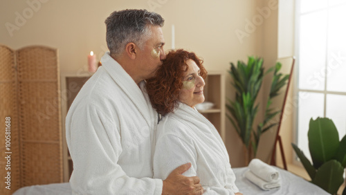 Middle-aged couple wearing white robes and eye masks embraces in a cozy wellness spa room, surrounded by plants and candles, enjoying a relaxing indoor experience.