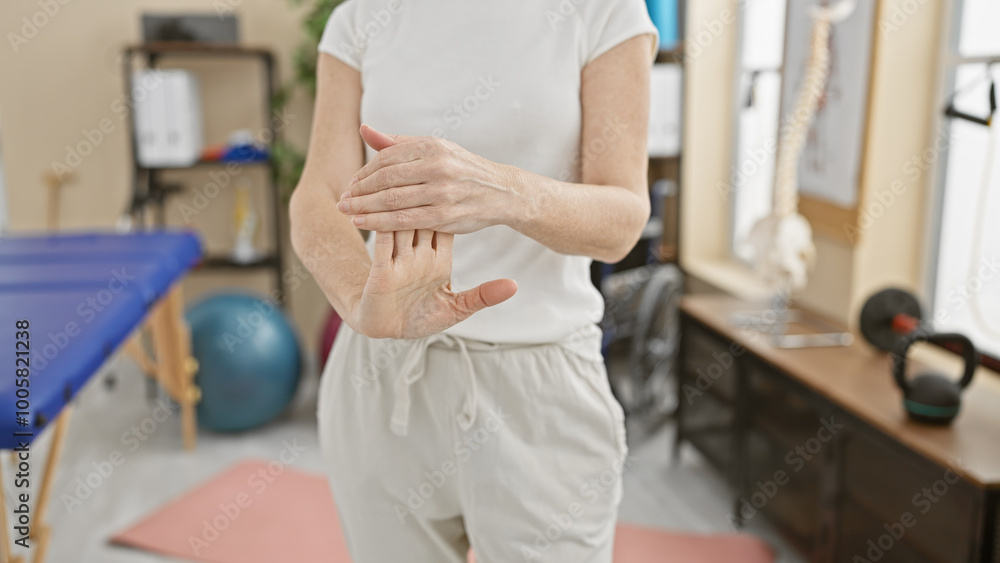 Fototapeta premium Mature woman exercises wrist in physiotherapy clinic with equipment background, portraying healthcare and rehabilitation.