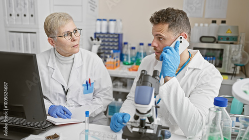 A female scientist and her male colleague in lab coats analyze samples in a laboratory while the man talks on the phone.