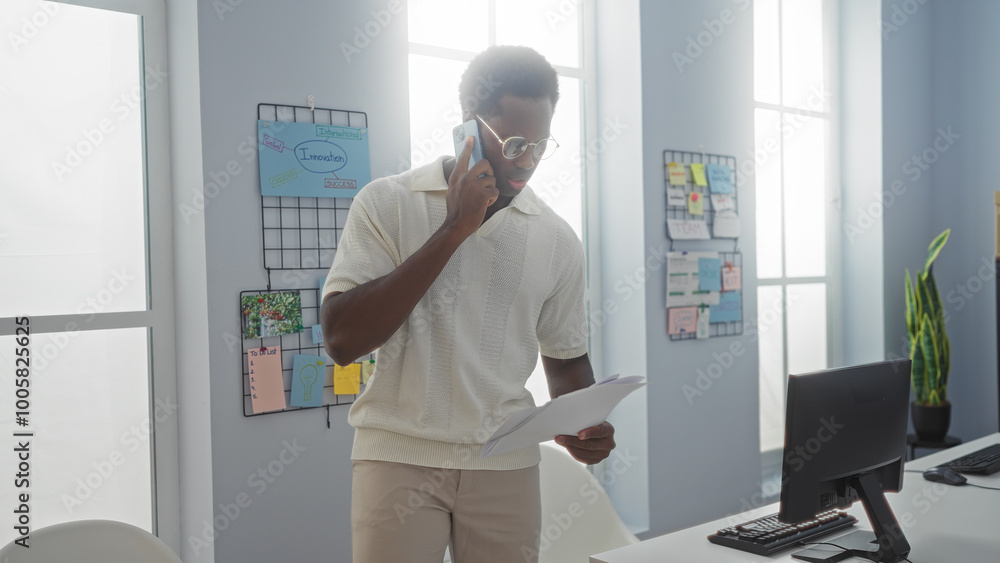 Young man talking on mobile phone while holding documents in modern office setting with bright light filtering through large windows and organized workspace. Young man talking on mobile phone while holding documents in modern office setting with bright light filtering through large windows and organized workspace.