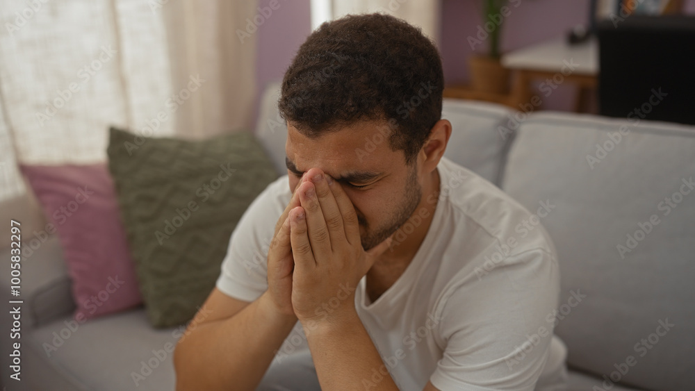 Young man sitting in a living room with hands covering his face, conveying a deep sense of stress or frustration, surrounded by cozy home decor. Young man sitting in a living room with hands covering his face, conveying a deep sense of stress or frustration, surrounded by cozy home decor.