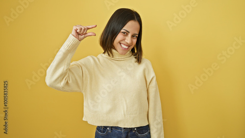 Smiling young hispanic woman making a small size gesture against a yellow background, portraying a casual and positive mood.