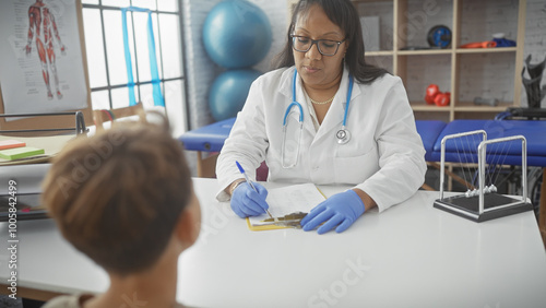 A woman doctor consults a boy patient in a clean, modern physiotherapy clinic room with rehabilitation equipment.