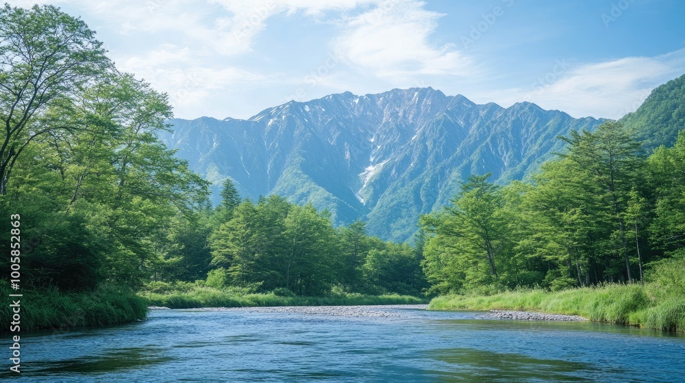 Fototapeta premium The peaceful Azusa River flowing through Kamikachi, framed by fresh green trees and the towering peaks of the Hotaka Mountains in the distance.