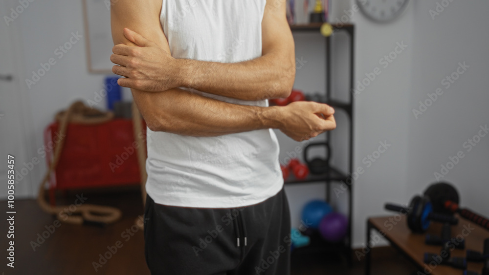Man experiencing arm pain in gymnasium, wearing white shirt, standing indoors surrounded by fitness equipment, highlighting a common injury associated with sports or exercise routines. Man experiencing arm pain in gymnasium, wearing white shirt, standing indoors surrounded by fitness equipment, highlighting a common injury associated with sports or exercise routines.