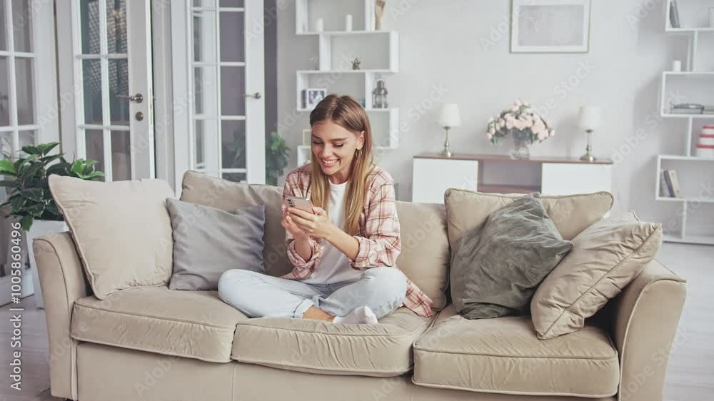 Young woman is using her smartphone while sitting on a sofa in her living room. She is wearing casual clothing and appears to be enjoying herself Young woman is using her smartphone while sitting on a sofa in her living room. She is wearing casual clothing and appears to be enjoying herself