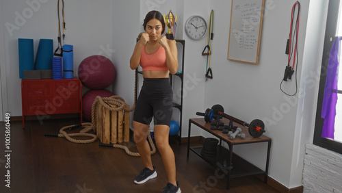 Young woman wearing workout clothes stands in a gym room with fitness equipment like ropes, dumbbells, and exercise balls, preparing for a workout session emphasizing health and wellness.