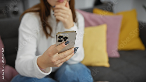 A thoughtful young woman holding a smartphone in a cozy living room