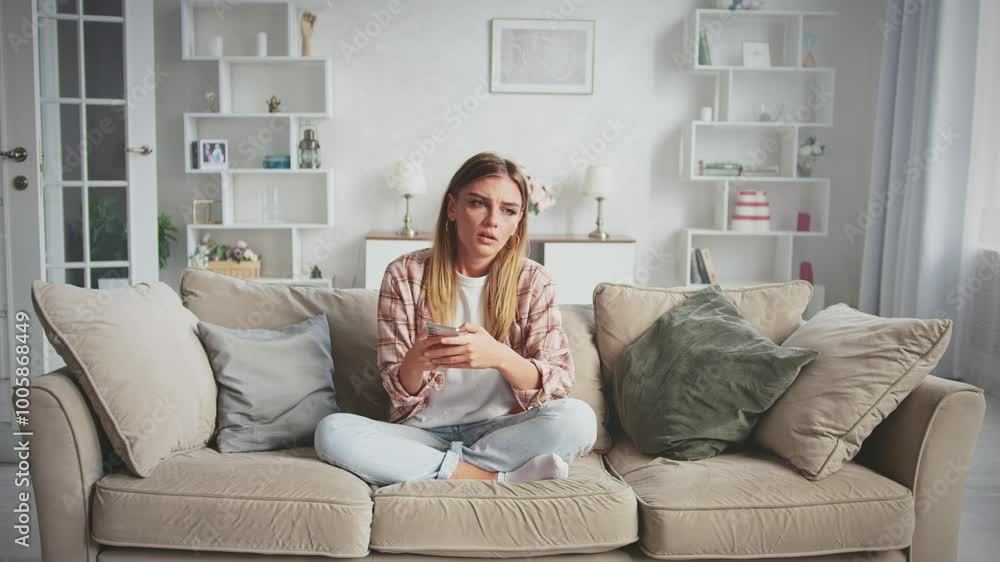 Young woman sits on a sofa in her living room, looking down at her smartphone with a sad expression. Feelings of loneliness and disappointment wash over her as she scrolls Young woman sits on a sofa in her living room, looking down at her smartphone with a sad expression. Feelings of loneliness and disappointment wash over her as she scrolls