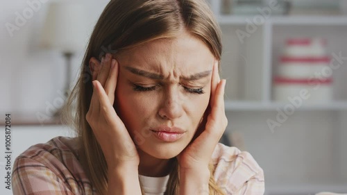 Young woman suffers from a strong headache or migraine at home. Massaging her temples and closing her eyes, she expresses pain and discomfort while feeling overwhelmed