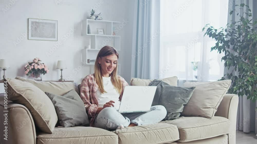 Young woman is sitting cross legged on a sofa in her living room, working on her laptop. She is smiling and appears to be enjoying her work Young woman is sitting cross legged on a sofa in her living room, working on her laptop. She is smiling and appears to be enjoying her work