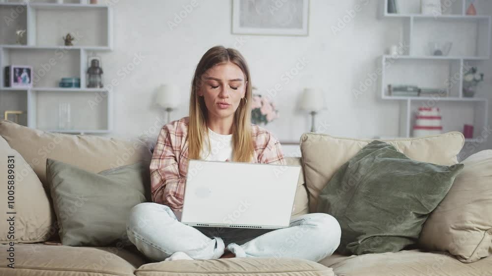 Young woman is using a laptop for a video call, sitting on a sofa in her living room. She is gesturing and looking at the screen Young woman is using a laptop for a video call, sitting on a sofa in her living room. She is gesturing and looking at the screen