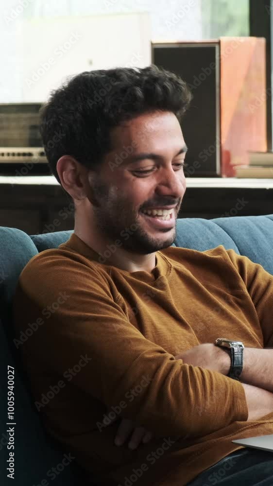 Positive Middle Eastern man spending leisure time with laptop, watching comedy movie and laughing, sitting on couch at home. Vertical video. Positive Middle Eastern man spending leisure time with laptop, watching comedy movie and laughing, sitting on couch at home. Vertical video.