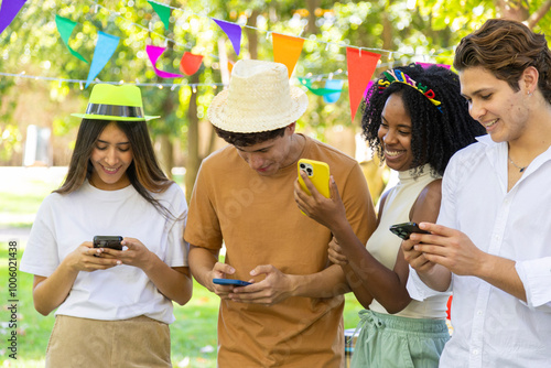 Four friends at an outdoor party wearing hats and using smartphones, smiling together.