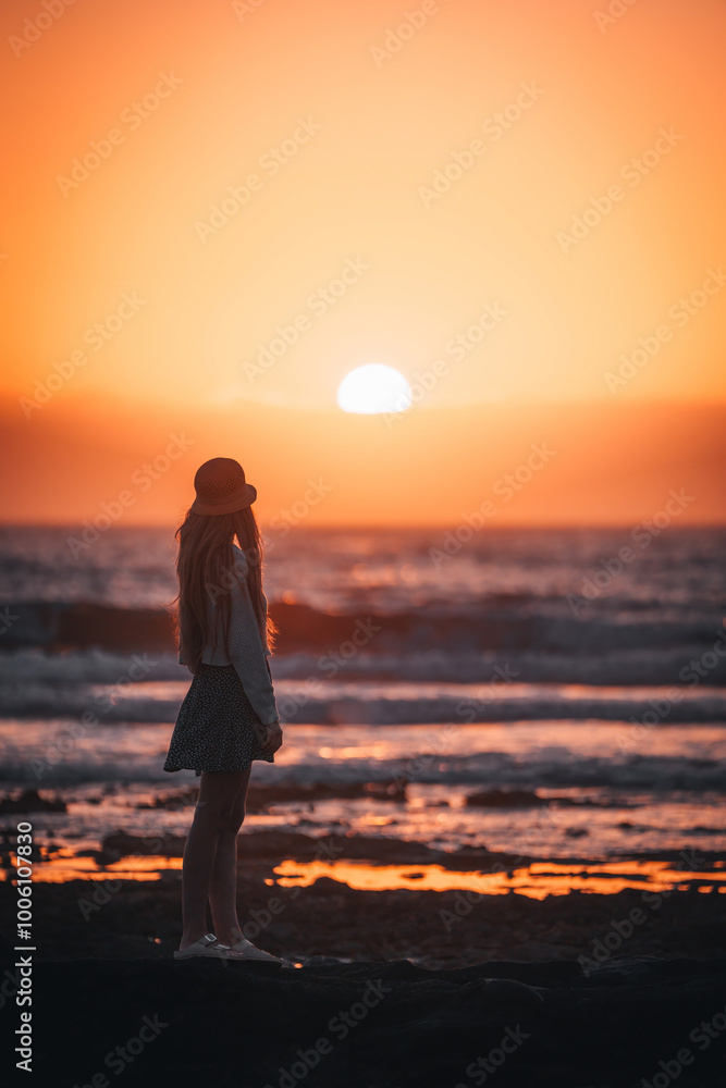 Fototapeta premium A woman stands on the beach, looking out at the sun as it sets