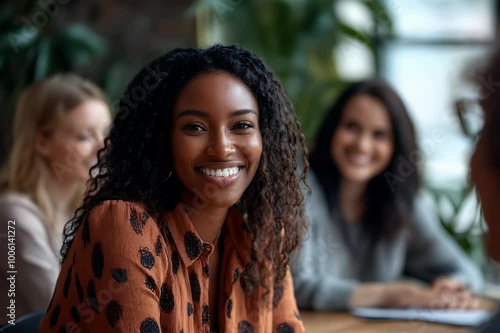 A curly-haired afro woman smiles at the camera, she is in an office. She is wearing a brown shirt.