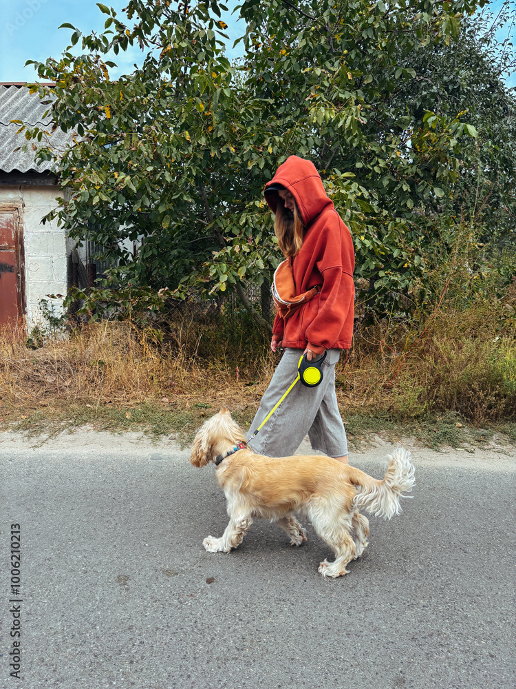 Side view of a woman walking her dog on a leash in casual attire. Streetwear fashion and outdoor lifestyle with a focus on pet care, ideal for content related to pets, health, or fashion.