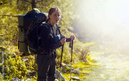 A beautiful smiling girl in the rays of the sun holds trekking poles and walks along a mountain trail. A young girl in thermal underwear and trekking pants with a backpack goes hiking in the forest.