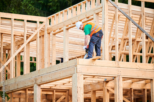 Carpenter using chainsaw for cutting wooden plank. Man worker building two-story wooden frame house. Concept of modern, environmentally-friendly construction.