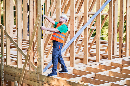 Carpenter constructing two-storey wooden frame house. Man inspects walls for levelness using spirit level, wearing protective overalls, helmet and vest. Concept of modern ecological construction.