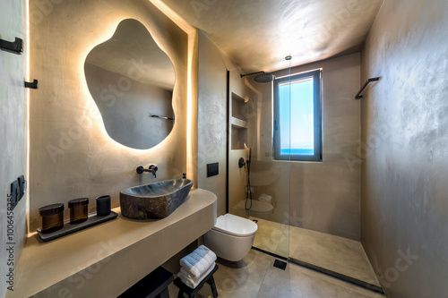 Modern bathroom interior design with cave-like in beige tones.  stone-like sink with natural finish, complemented by unusually shaped mirror with stylish backlighting. Spacious walk-in shower .