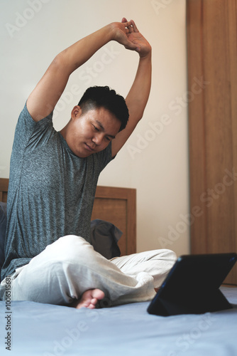 An asian young man sitting on the bed and stretch oneself in the bed room. Relaxation from reading a book. E-learning and and stretching to get relaxed, Education and learning concept.