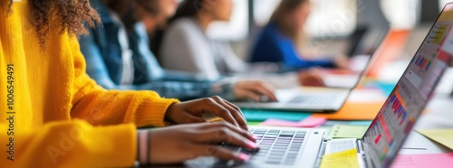 Close-up of young people working together on a laptop computer with colorful papers and pens at an office desk.
