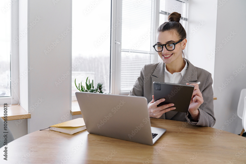 A Young Woman With Glasses Works on a Tablet While Sitting at a Desk With a Laptop in a Bright, Modern Office Setting A Young Woman With Glasses Works on a Tablet While Sitting at a Desk With a Laptop in a Bright, Modern Office Setting