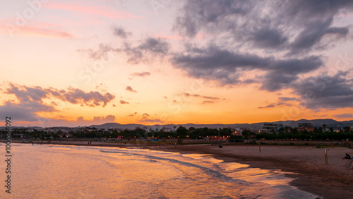 Paisaje al atardecer con cielo nublado en tonos rosas y anaranjados reflejados sobre la playa y el mar tranquilo, con silueta de ciudad al fondo photo