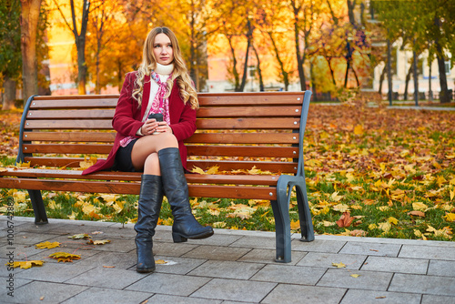 A beautiful woman in a red coat sits on a bench in an autumn park. She is wearing black boots and a scarf.