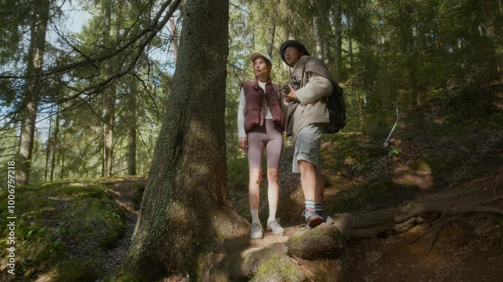 Full shot of interracial couple of hikers enjoying lush forest setting while standing on trail beside tall green tree and having conversation, looking straight ahead at picturesque landscape Full shot of interracial couple of hikers enjoying lush forest setting while standing on trail beside tall green tree and having conversation, looking straight ahead at picturesque landscape