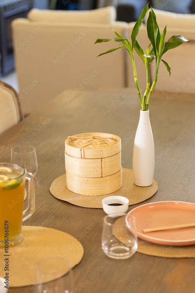 Fototapeta premium Dining table with bamboo steamer, vase with bamboo plant, and lemonade pitcher