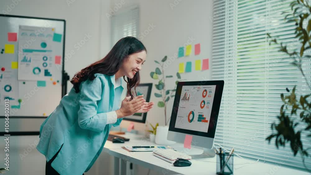 Asian businesswoman in a blue suit is clapping her hands in front of a computer screen with graphs. She is standing up in her office and seems very happy about the results Asian businesswoman in a blue suit is clapping her hands in front of a computer screen with graphs. She is standing up in her office and seems very happy about the results