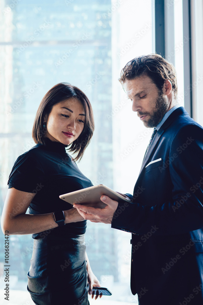 © BullRun - Male and female lawyers checking information on touch pad standing near skyscraper window in office, successful diverse employees reading financial news on digital tablet, colleagues collaborating