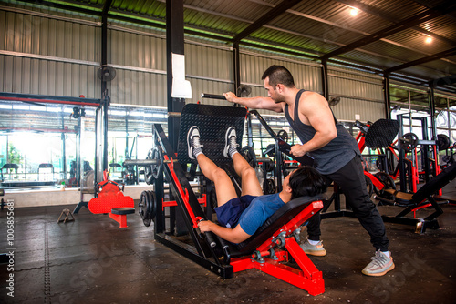 A male trainer is helping his trainee use a leg press machine by pushing the handles of the machine.