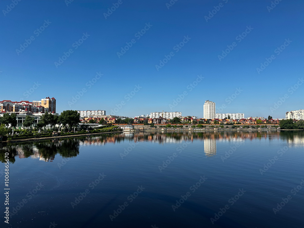 Naklejka premium clear, calm view of a city reflected perfectly in the still waters of a lake. The skyline includes residential buildings, trees, and a bright blue sky, creating a serene and picturesque scene.